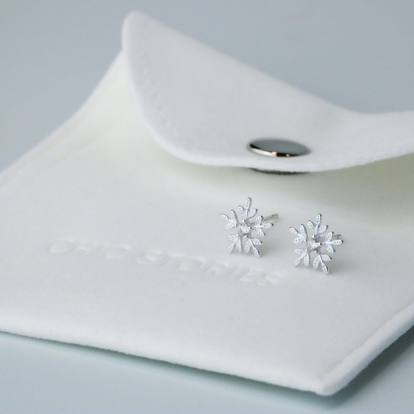 Silver snowflake shape stud earrings with a white crystal detail in the centre. They are sitting on top of a white felt jewellery pouch, embossed with 'Chic Stories".