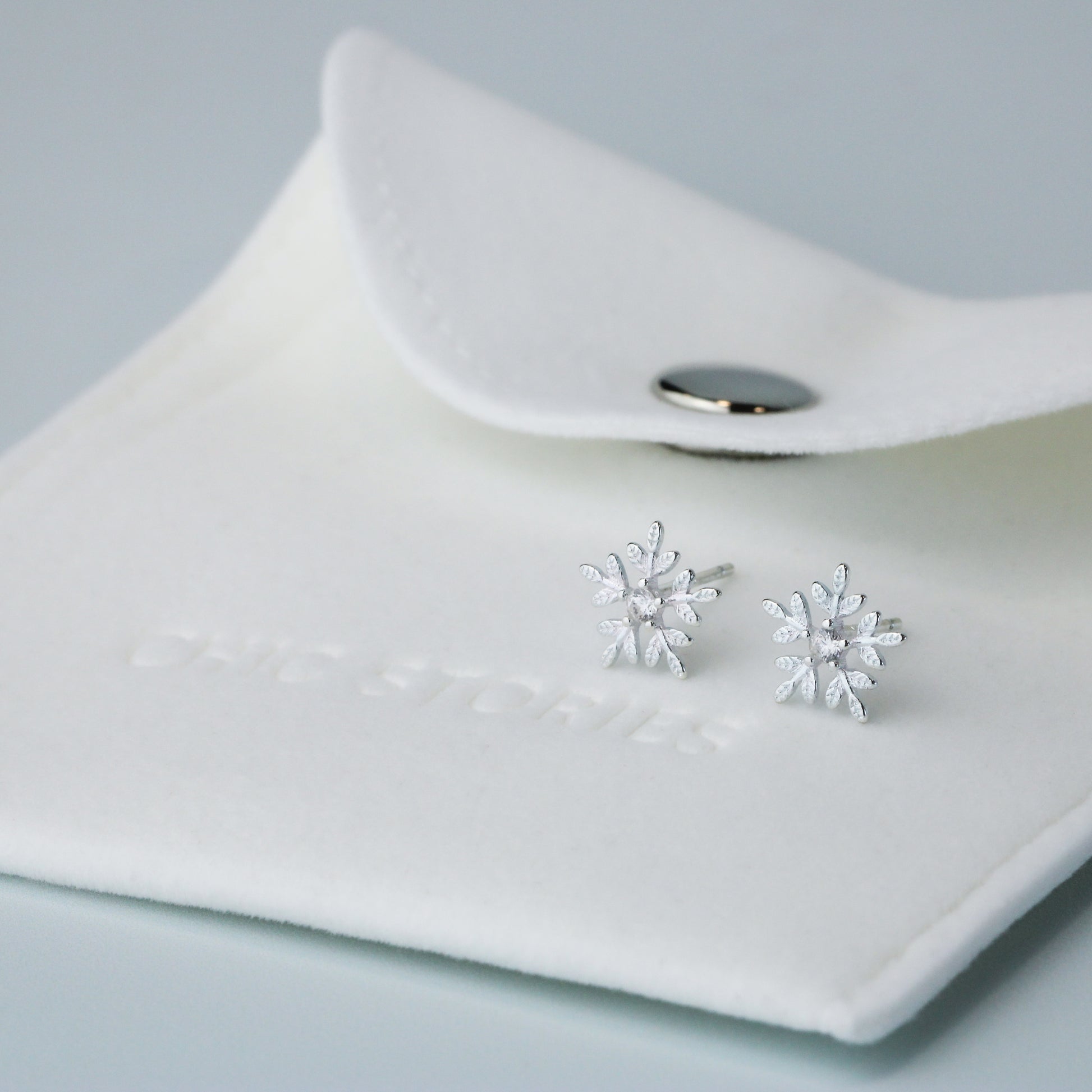Silver snowflake shape stud earrings with a white crystal detail in the centre. They are sitting on top of a white felt jewellery pouch, embossed with 'Chic Stories".