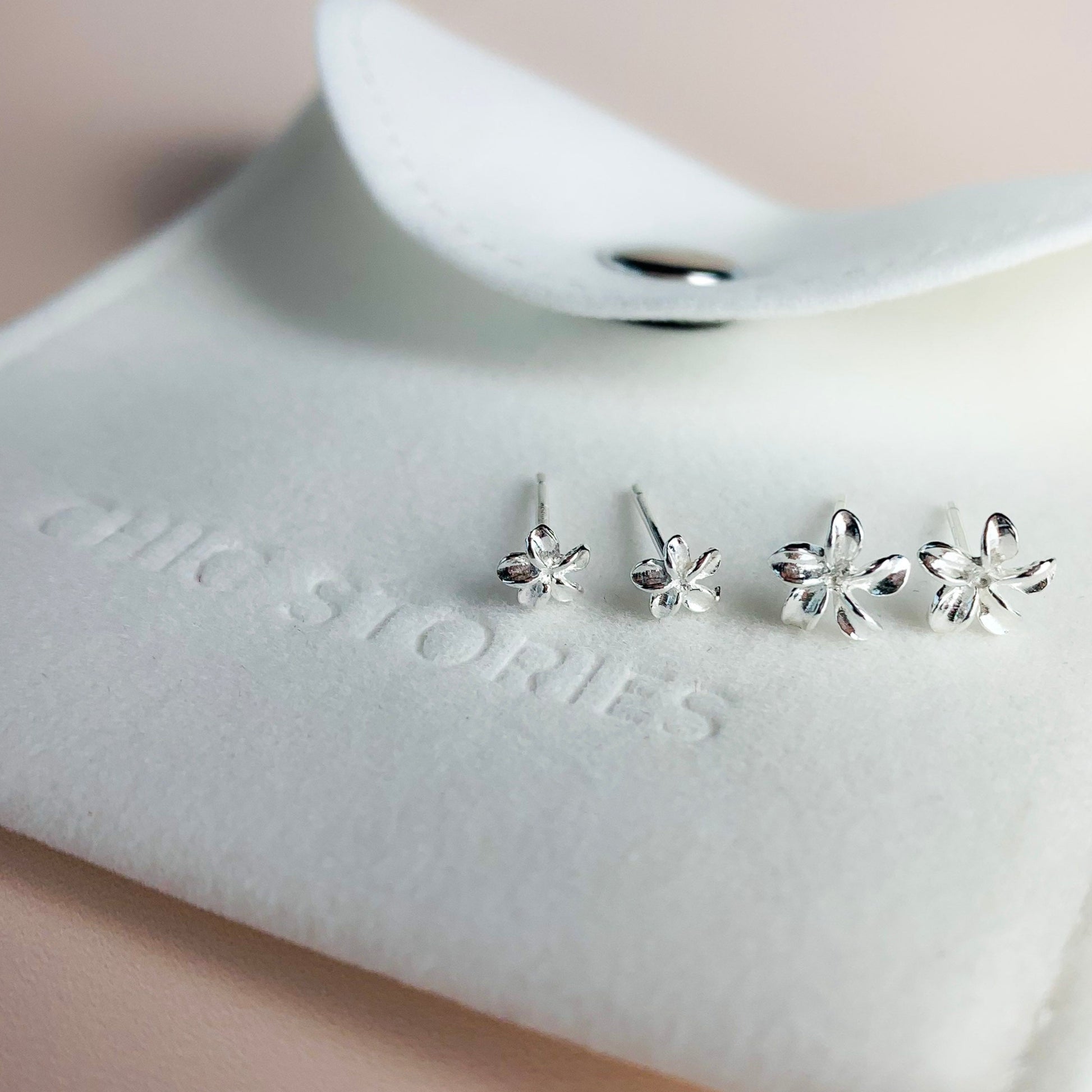 Two pairs of sterling silver flower stud earrings, in two different size options. They are sitting on a white felt jewellery pouch, embossed with the words Chic Stories