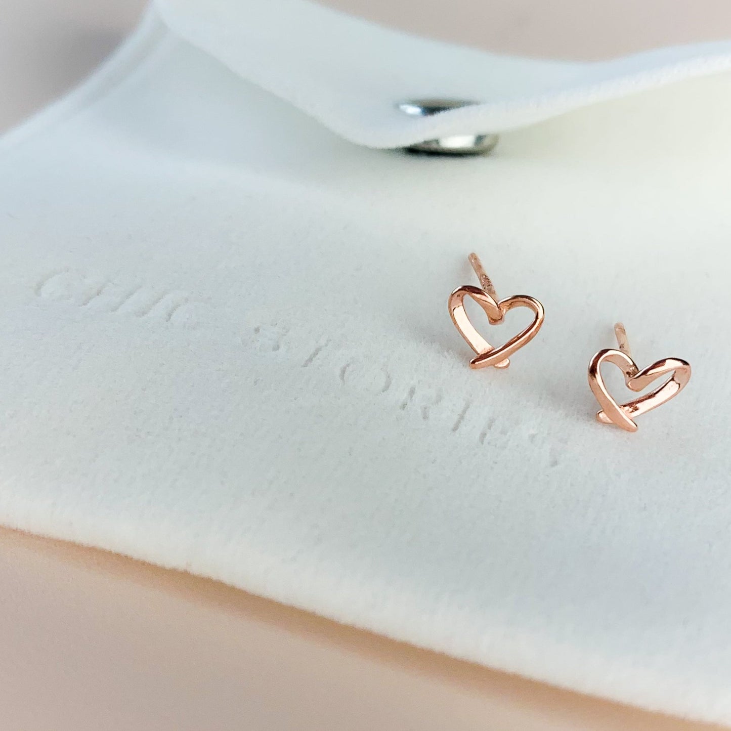 Rose gold heart stud earrings, sitting on a white felt jewellery pouch, embossed with 'Chic Stories"