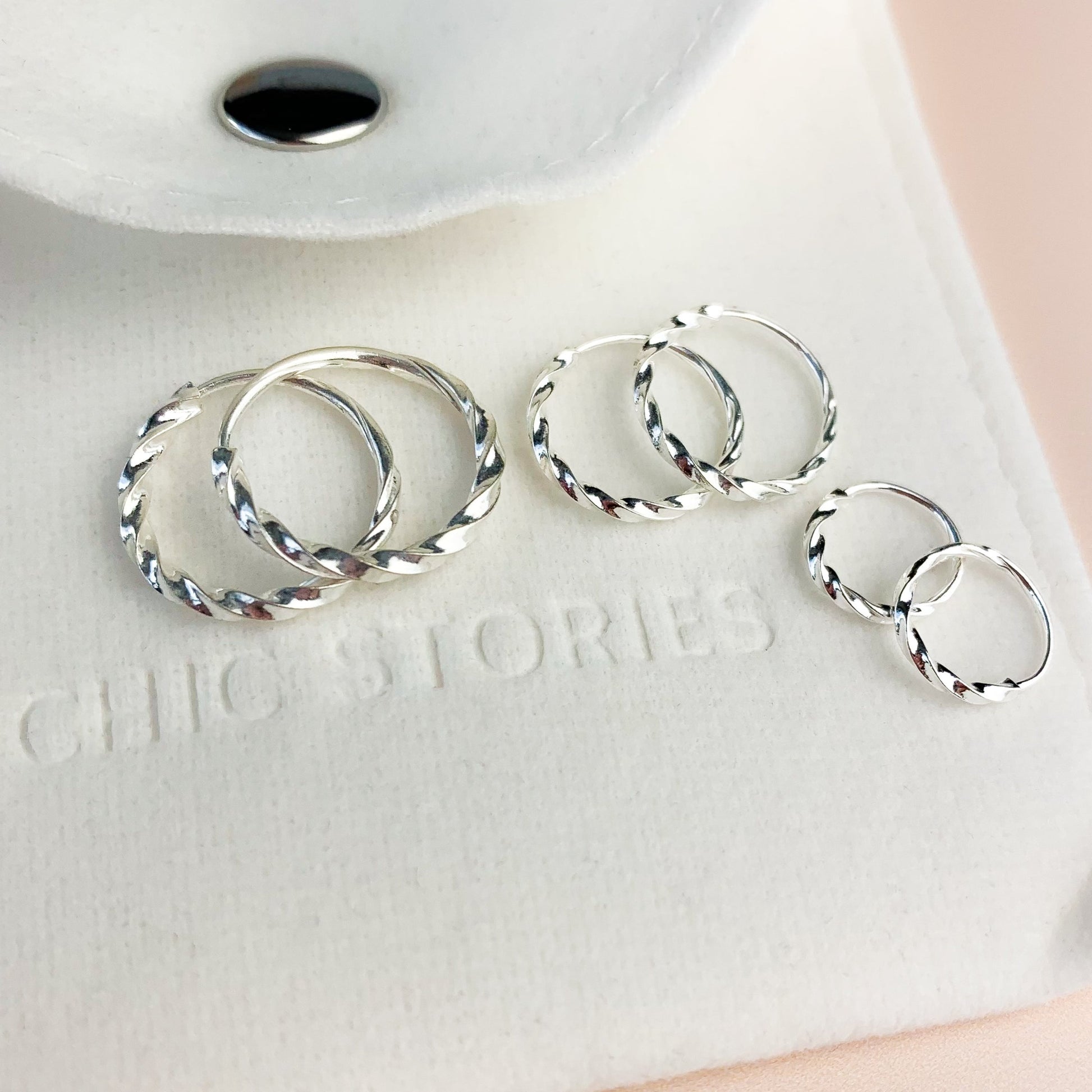 Twisted silver hoop earrings with a sleeper fastening, in three sizes. They sit on a white felt jewellery pouch, embossed with 'Chic Stories'